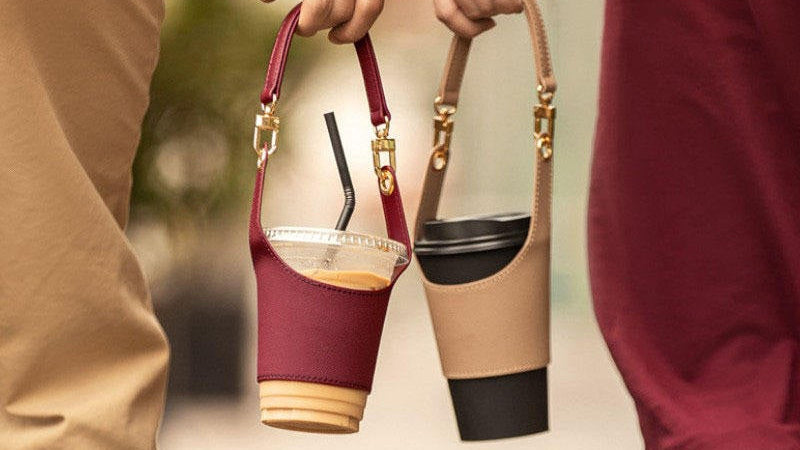 Two people holding reusable insulated cup sleeves in a close-up lifestyle shot. One sleeve is burgundy with gold hardware and a clear cup insert, and the other is beige with gold hardware and a black cup insert.
