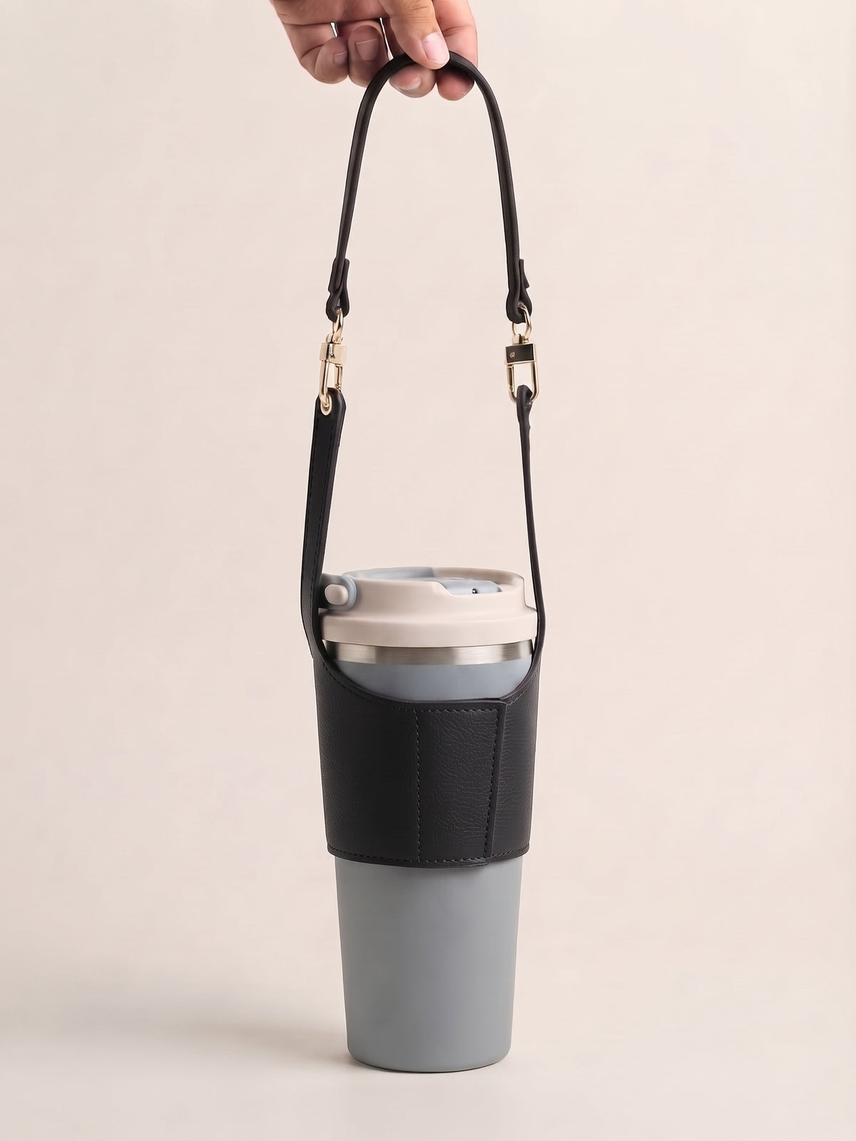 A gray insulated travel tumbler with a black sleeve and matching black shoulder strap, held by a hand against a plain beige background.