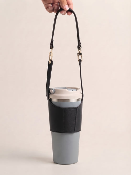A gray insulated travel tumbler with a black sleeve and matching black shoulder strap, held by a hand against a plain beige background.